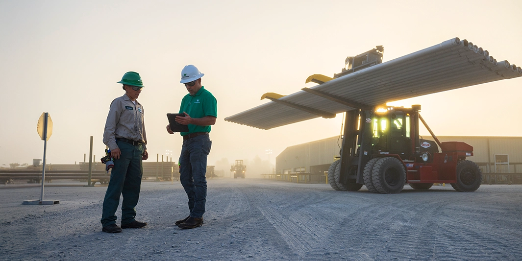 Two Workstrings workers looking over inventory on a tablet with a large forklift holding drill pipes in the background